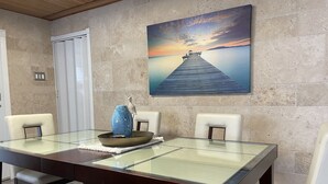 A dining area with a glass table, two chairs, a vase, and a painting of a sunset over water. - Casa Oceana Minutes to Seven Seas Beach + Bio Bay + Marinas (Fajardo)