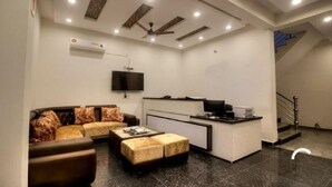 A modern living room with a sofa, ottoman, and a small coffee table. There is a flat-screen TV mounted on the wall and a desk with a computer and printer. The room has a ceiling fan and recessed lighting.