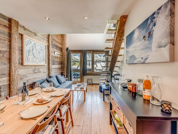 Un coin repas chaleureux avec une table en bois dressée pour deux personnes, un canapé et une vue sur les montagnes grâce à de grandes fenêtres.