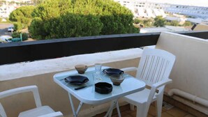 Un petit balcon avec une table dressée pour deux personnes, deux chaises en plastique blanches et une vue sur un paysage urbain, avec des arbres et des immeubles.