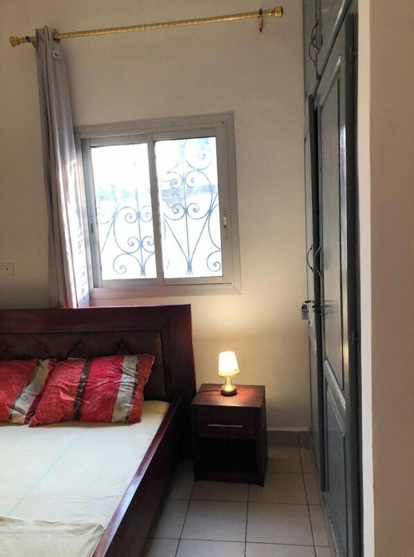Une chambre à coucher avec un lit, une table de chevet avec une lampe, une fenêtre ornée de barreaux en fer décoratifs et une porte.