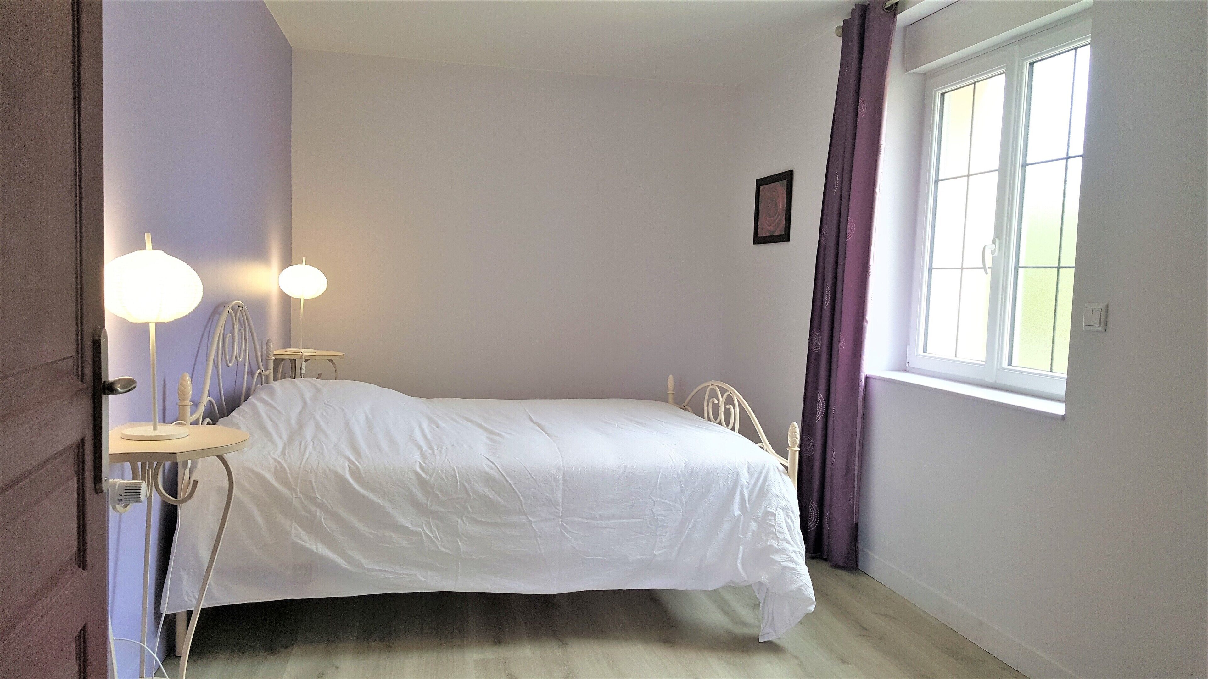 A bedroom with a single bed, bedside lamps, a window with curtains, and a small table with a lamp.