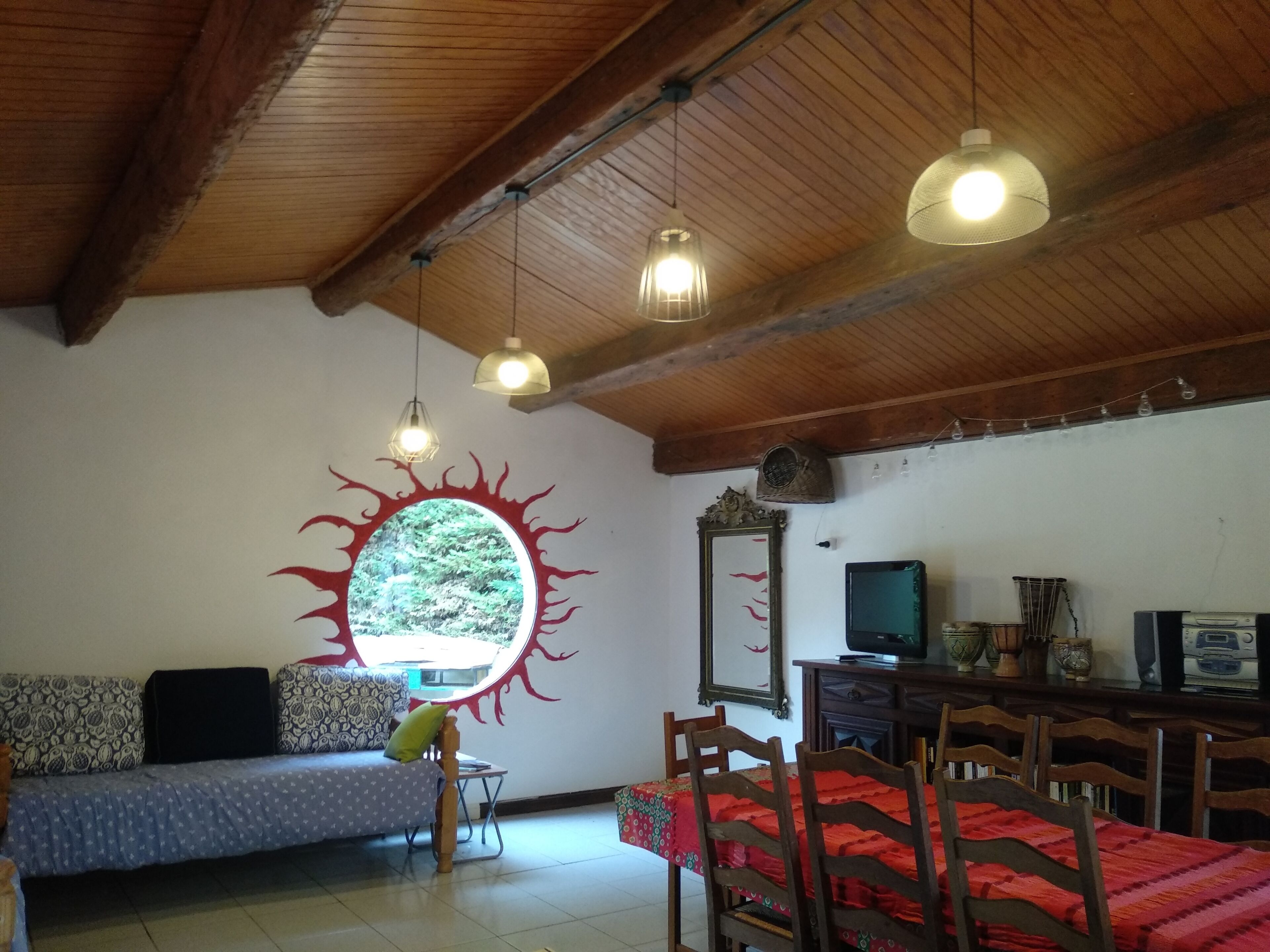 Een gezellige woonkamer met een houten plafond, een ronde wanddecoratie, een bank, een eettafel met stoelen en een televisie.