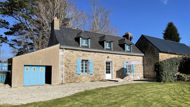 A stone house with a blue garage door, a stone chimney, and a small porch with a table and chairs. - QUIET STONE HOUSE 3KMS FROM THE BEACH (La Foret-Fouesnant)