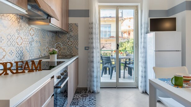 A modern kitchen with patterned tiles, a wooden cabinet, a countertop with a 'CREAM' sign, a sliding glass door to a balcony, and a dining table with chairs.