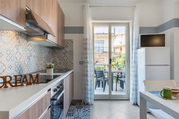 Une cuisine moderne avec des carreaux décoratifs, un meuble en bois, un plan de travail orné d’une inscription « CREAM », une porte-fenêtre coulissante donnant sur un balcon et une table à manger avec des chaises.