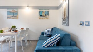 A modern living room with a blue sofa, white dining table, and chairs, a small plant, and two wall-mounted paintings.