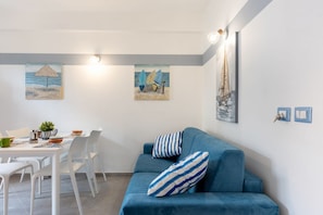 A modern living room with a blue sofa, white dining table, and chairs, a small plant, and two wall-mounted paintings.