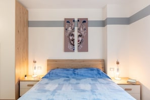 A bedroom with a wooden headboard, a bed with a blue textured bedspread, two bedside tables with lamps, and a wall-mounted artwork.