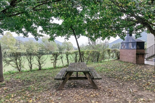A picnic table under a tree with a brick barbecue grill in the background.