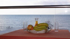 Una mesa con frutero que contiene plátanos, manzanas y un vaso de jugo, con vista al mar.