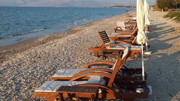 Una playa con sillones de madera y sombrillas, arena de guijarros y vistas al mar y montañas a la distancia.