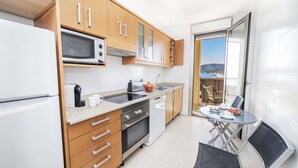 A modern kitchen with wooden cabinets, a microwave, and a sink. There is a dining area with a table and chairs, and a view of the sea through an open door. - Apartamento Bellavista Puerto 2A (Cala Ratjada)