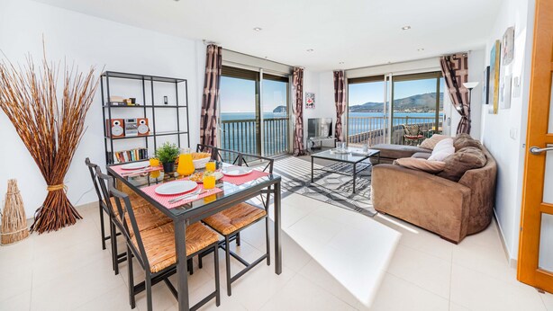A modern living room with a dining area, large windows, and a balcony overlooking a scenic view. - Apartamento Bellavista Puerto 2A (Cala Ratjada)