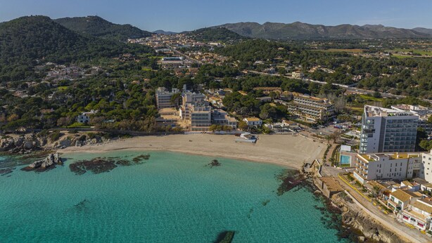 An aerial view of a coastal town with a beach, buildings, and mountains in the background. - Apartamento Bellavista Puerto 2B (Cala Ratjada)