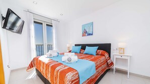 A modern bedroom with a large bed, a flat-screen TV mounted on the wall, a painting above the bed, and a small bedside table with a lamp. - Apartamento Bellavista Puerto 2B (Cala Ratjada)