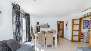A modern living room with a grey sofa, a dining area with a white table and chairs, and a kitchen area with wooden cabinets and a hanging plant. - Apartamento Bellavista Puerto 2B (Cala Ratjada)