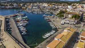 A marina with numerous boats docked, surrounded by residential buildings and a clear view of the mountains in the background. - Apartamento Bellavista Puerto 2B (Cala Ratjada)