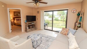 A living room with a sofa, a TV on a wooden stand, a ceiling fan, and a view of a garden through a sliding glass door.