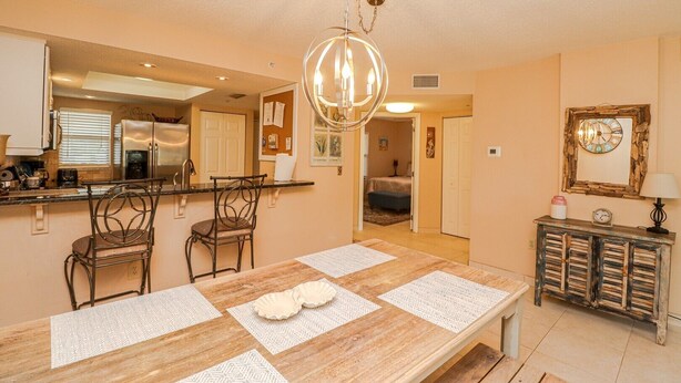 A dining area with a wooden table, chairs, and a chandelier. There is a kitchen area with a refrigerator and a sink.