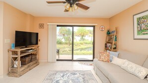 A living room with a television on a wooden stand, a ceiling fan, a sofa with patterned cushions, and a view of a garden through a sliding glass door.
