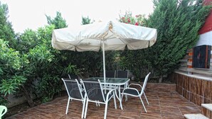 An outdoor patio with a table and chairs under a white umbrella, surrounded by greenery and a tiled floor. - Villa Ariadni Crete (Agios Vasileios)