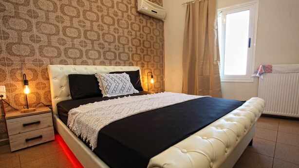 A bedroom with a bed, nightstands, a wall-mounted air conditioner, a window with curtains, and a radiator. - Villa Ariadni Crete (Agios Vasileios)