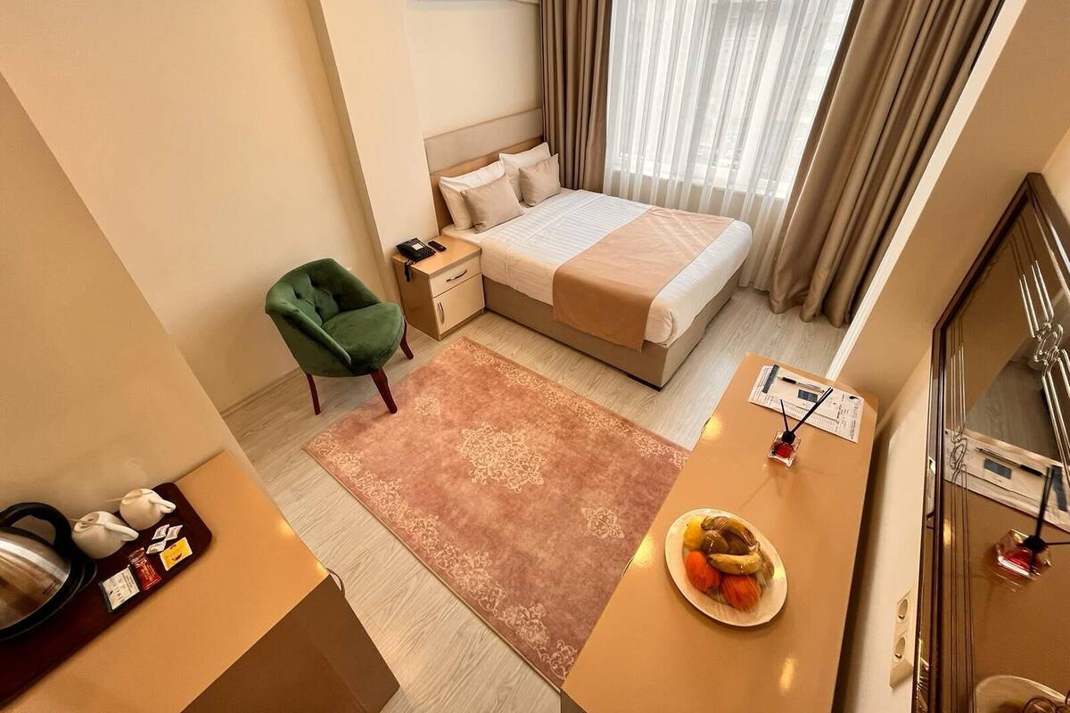 A hotel room with a bed, a desk with a plate of fruit, a chair, a bedside table, and a window with curtains.