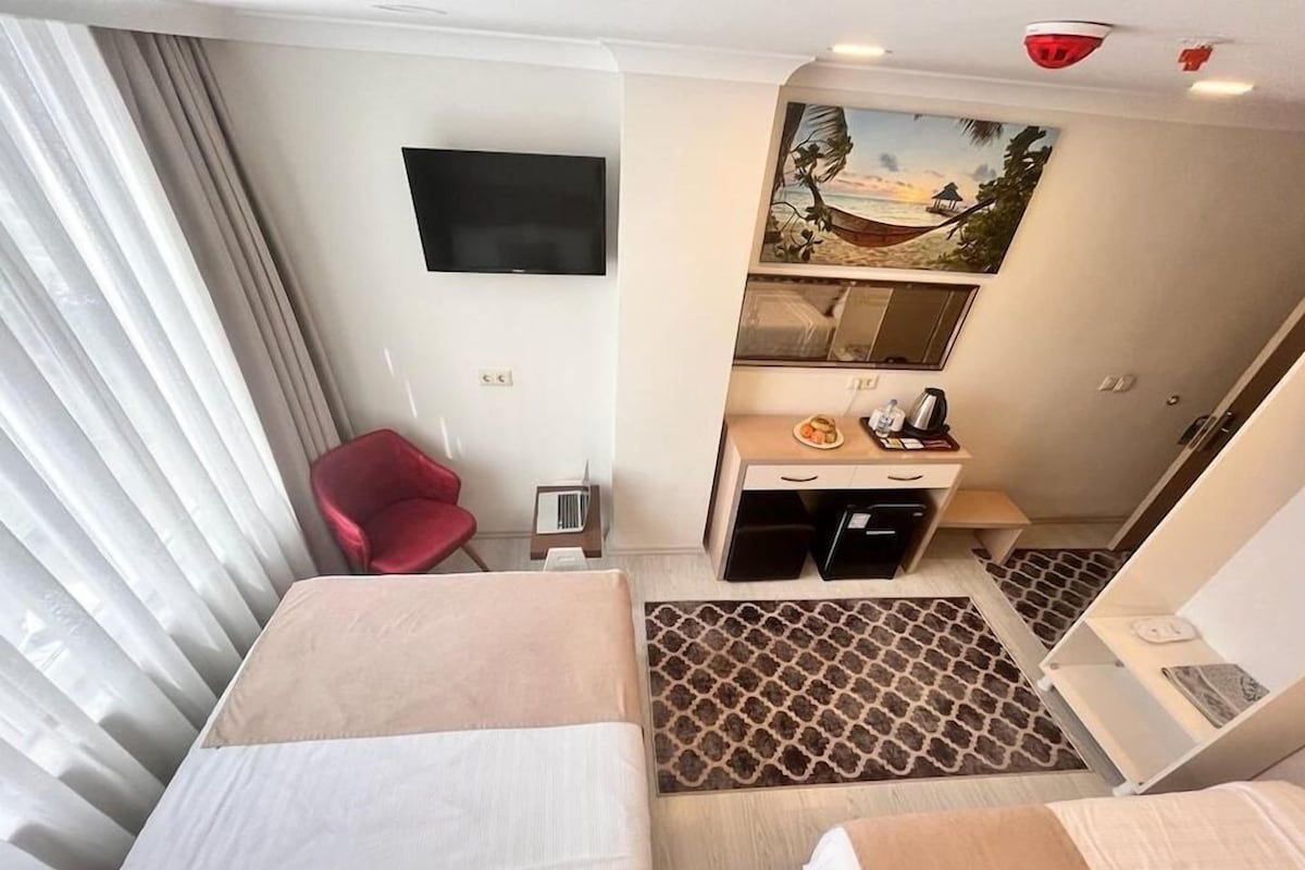 Ett hotellrum med två sängar, en tv som är monterad på väggen, ett skrivbord med en stol och en tavla som föreställer en strandvy.