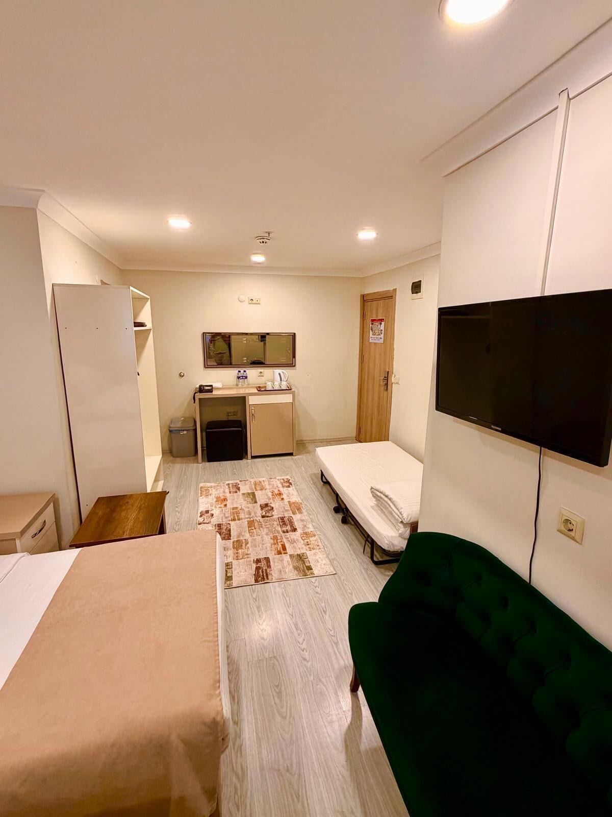 A hotel room with a bed, a green sofa, a TV, a desk, a mirror, and a small kitchenette.