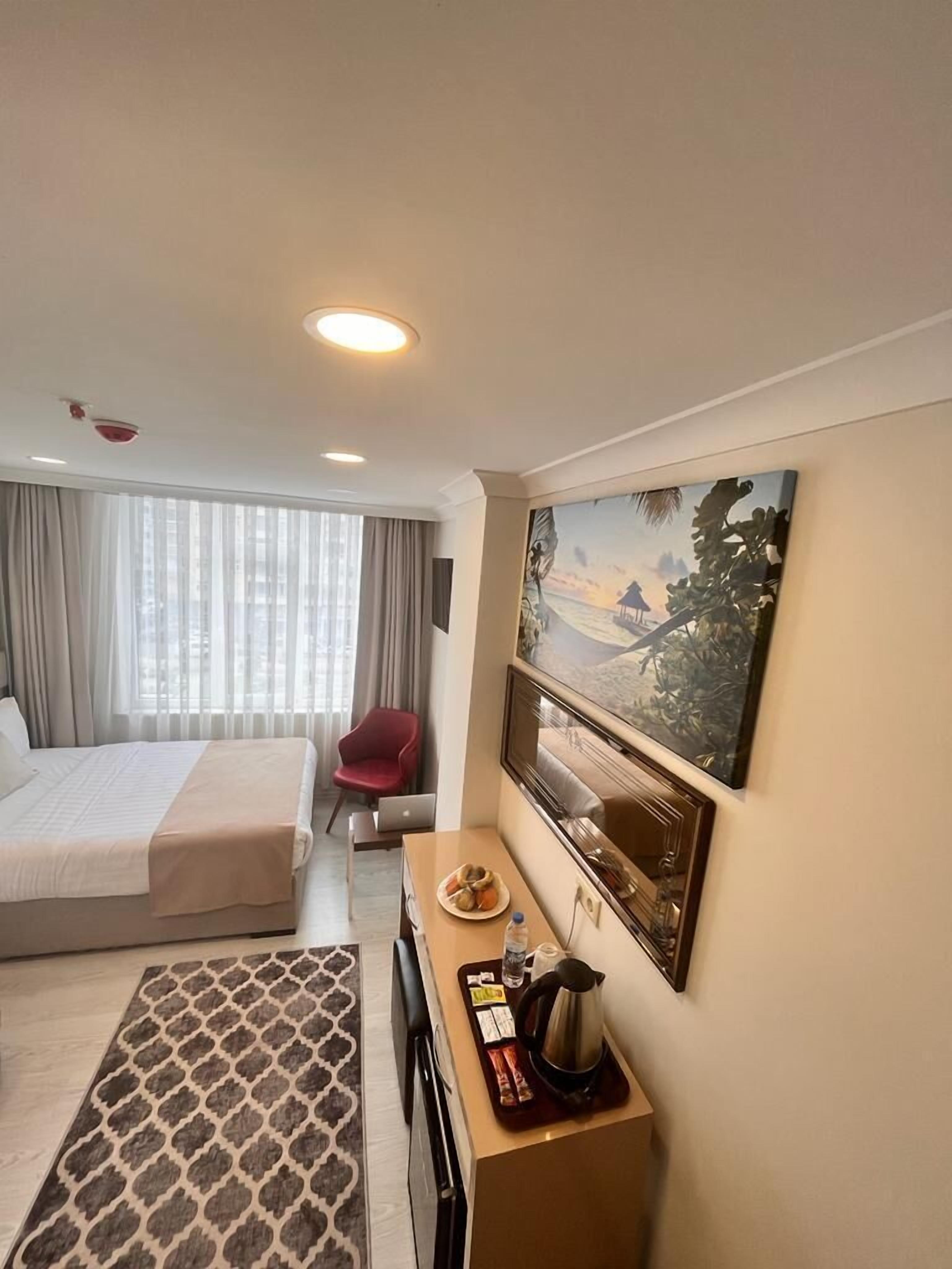 A hotel room with a bed, a desk with a coffee maker, a chair, and a painting on the wall.