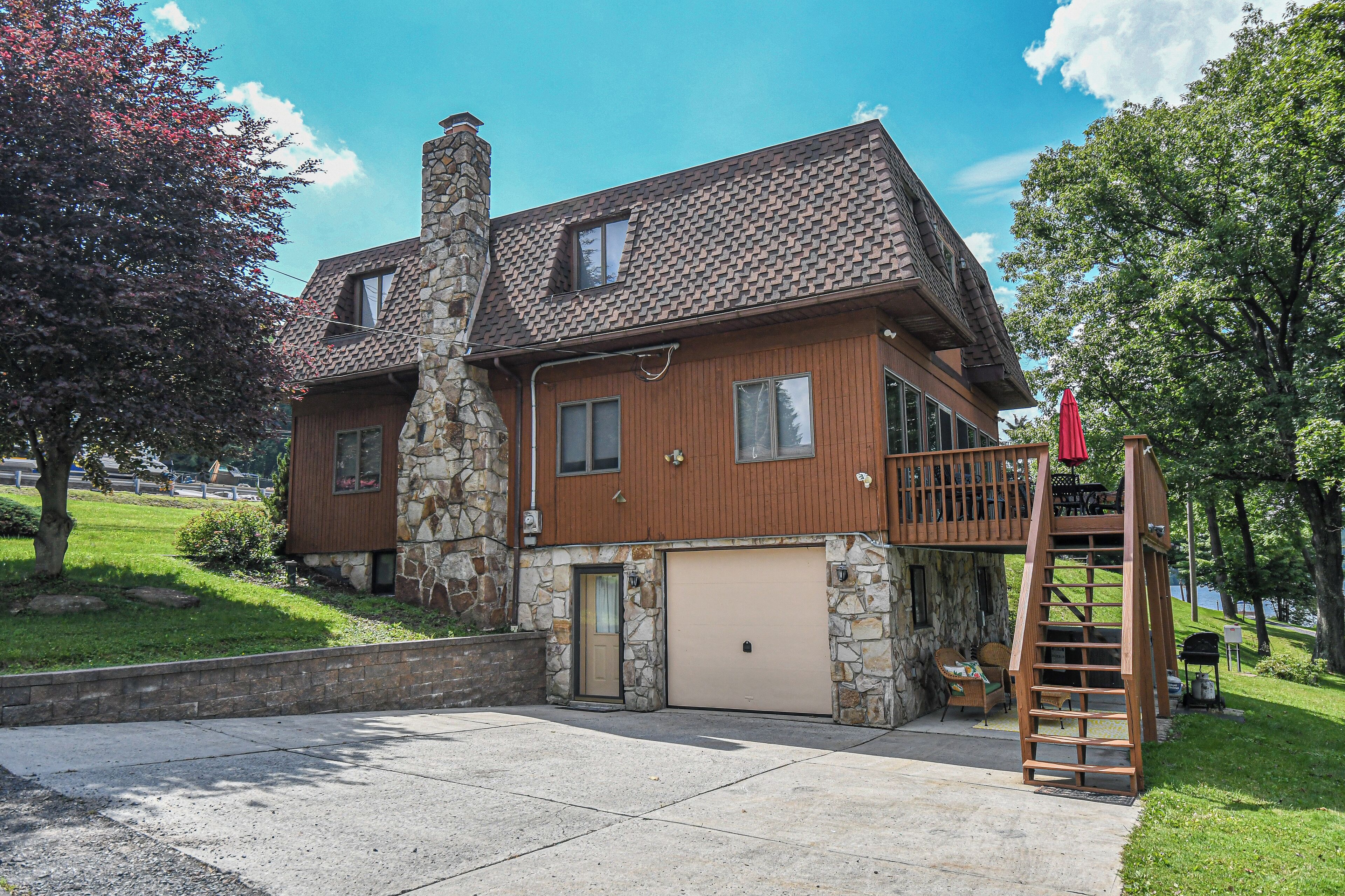 Une maison à deux étages avec une cheminée en pierre, un garage et une terrasse en bois surmontée d’un parasol rouge.