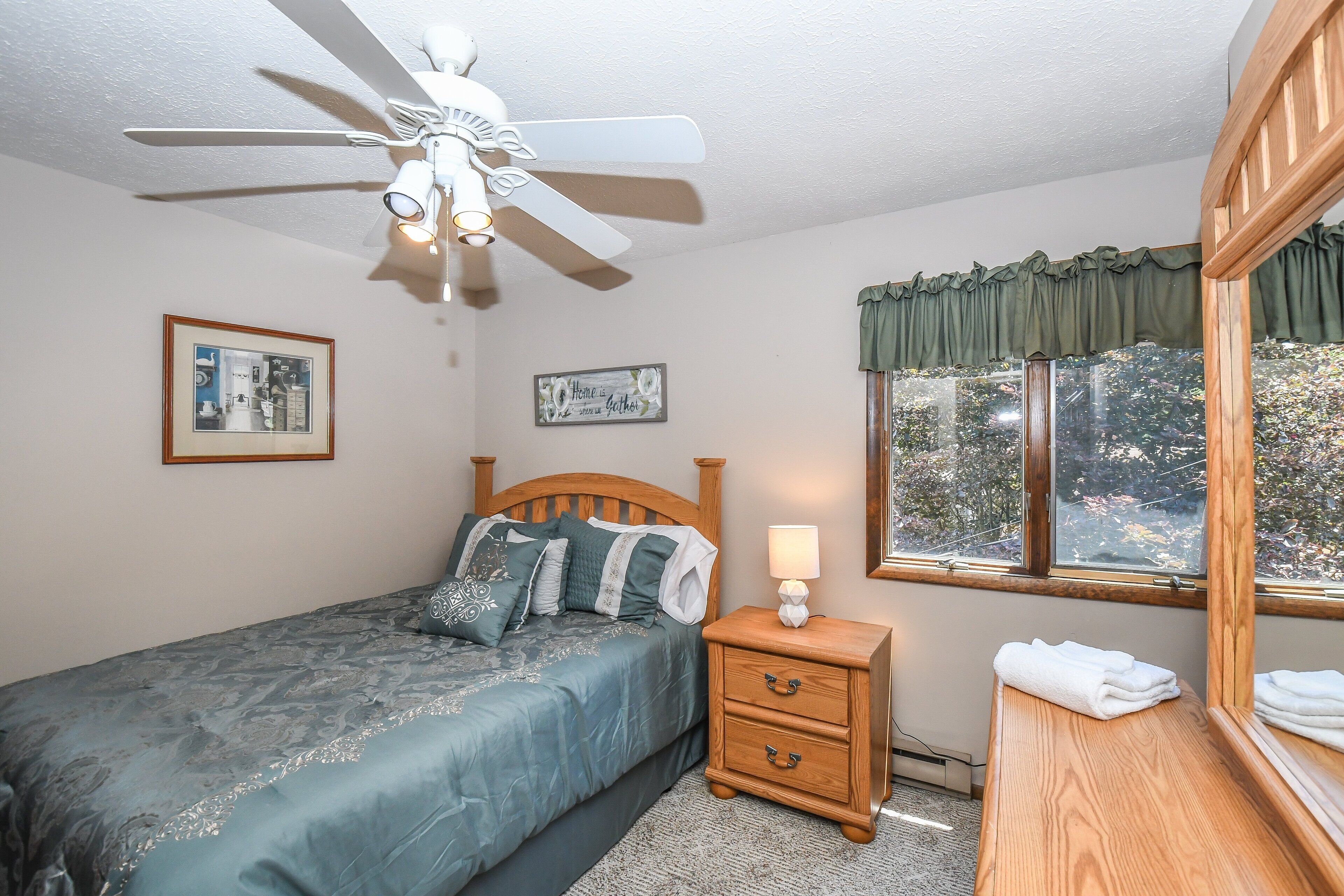 A bedroom with a bed, a nightstand, a ceiling fan, a window with curtains, and two wall decorations.