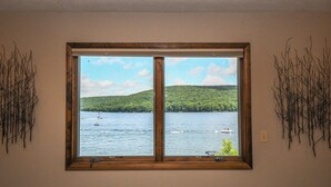 A room with a large window offering a view of a lake with boats and a green hill. - Canoe Cottage by Taylor Made Deep Creek (Oakland)