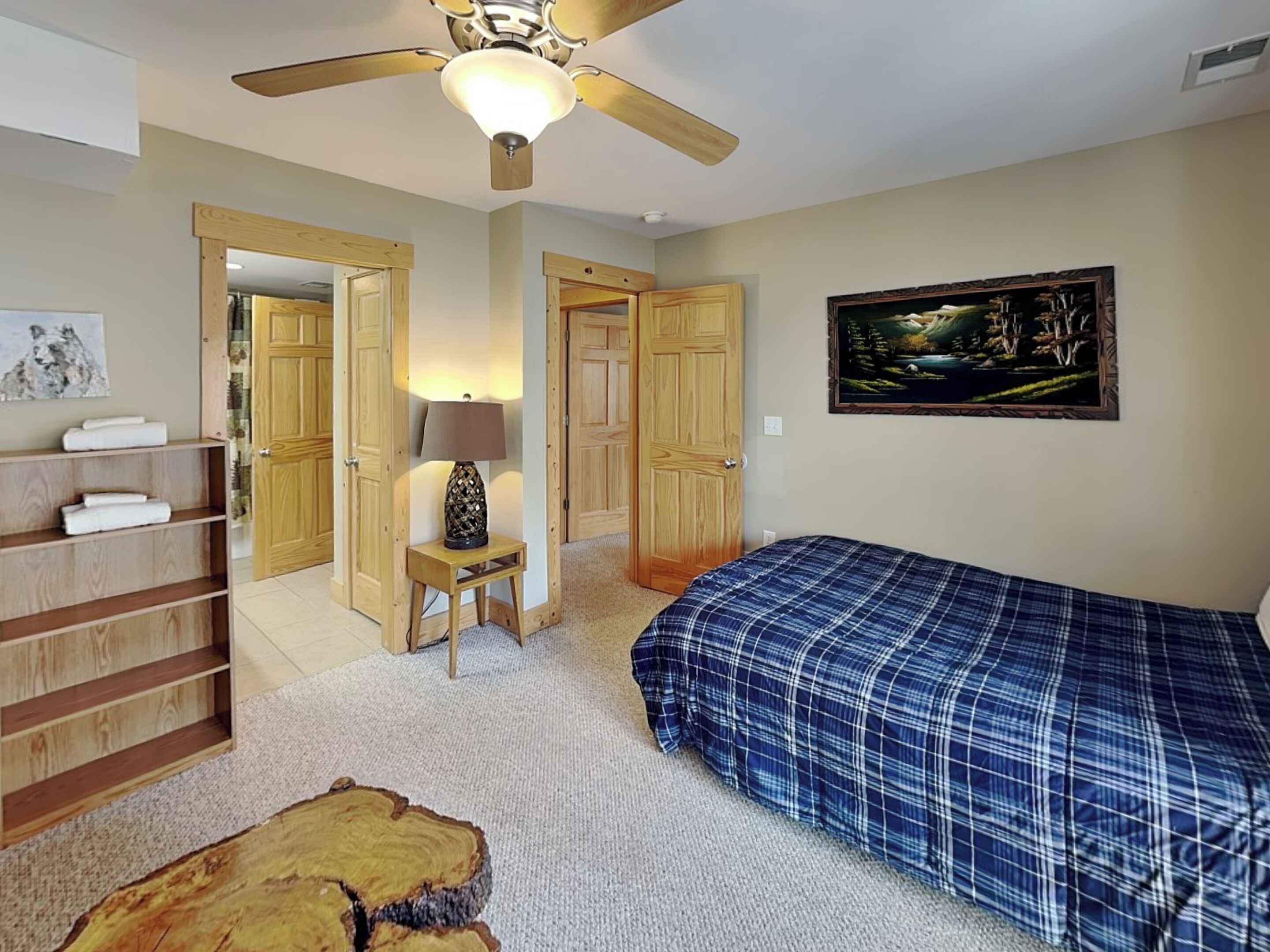 A bedroom with a bed, a ceiling fan, a wooden wardrobe, a nightstand with a lamp, a painting on the wall, and a wooden shelf with towels.