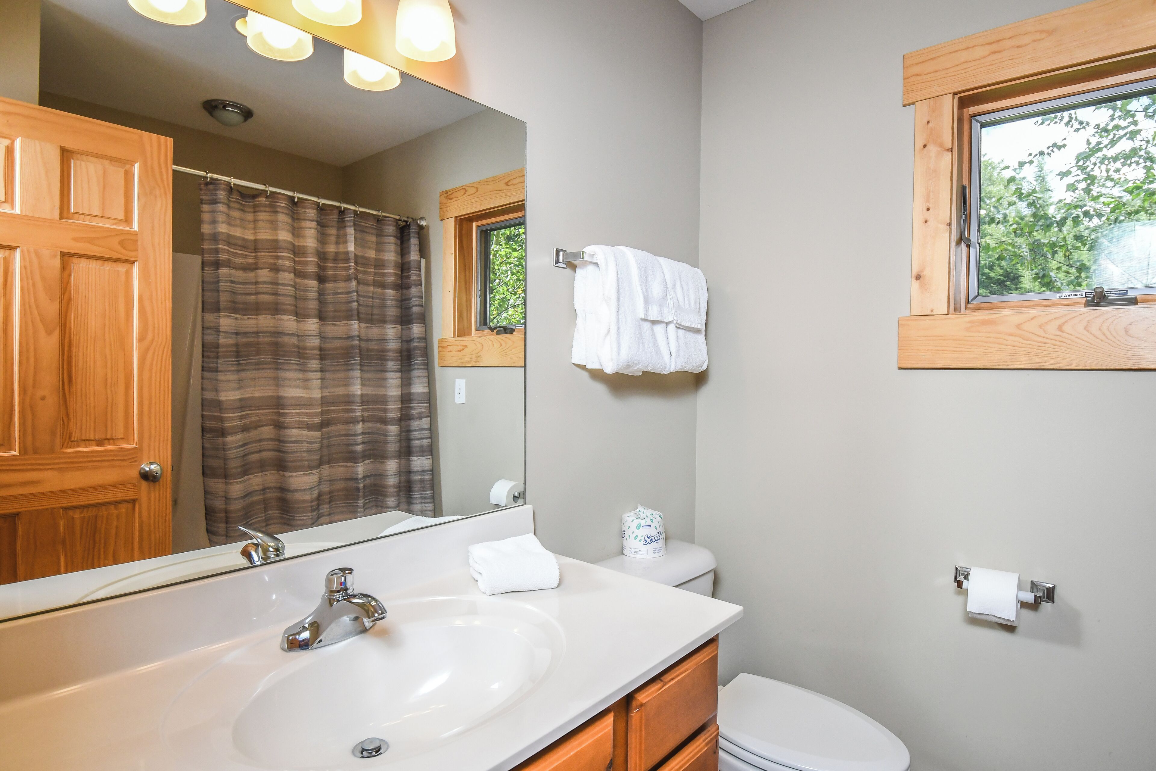 Une salle de bain avec une porte en bois, un grand miroir, un lavabo blanc, des toilettes et une fenêtre donnant sur des arbres.