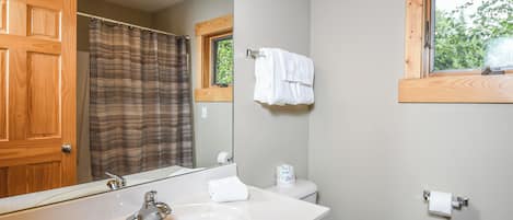 Ein Badezimmer mit Holztür, einem großen Spiegel, einem weißen Waschbecken, einer Toilette und einem Fenster mit Blick auf Bäume.