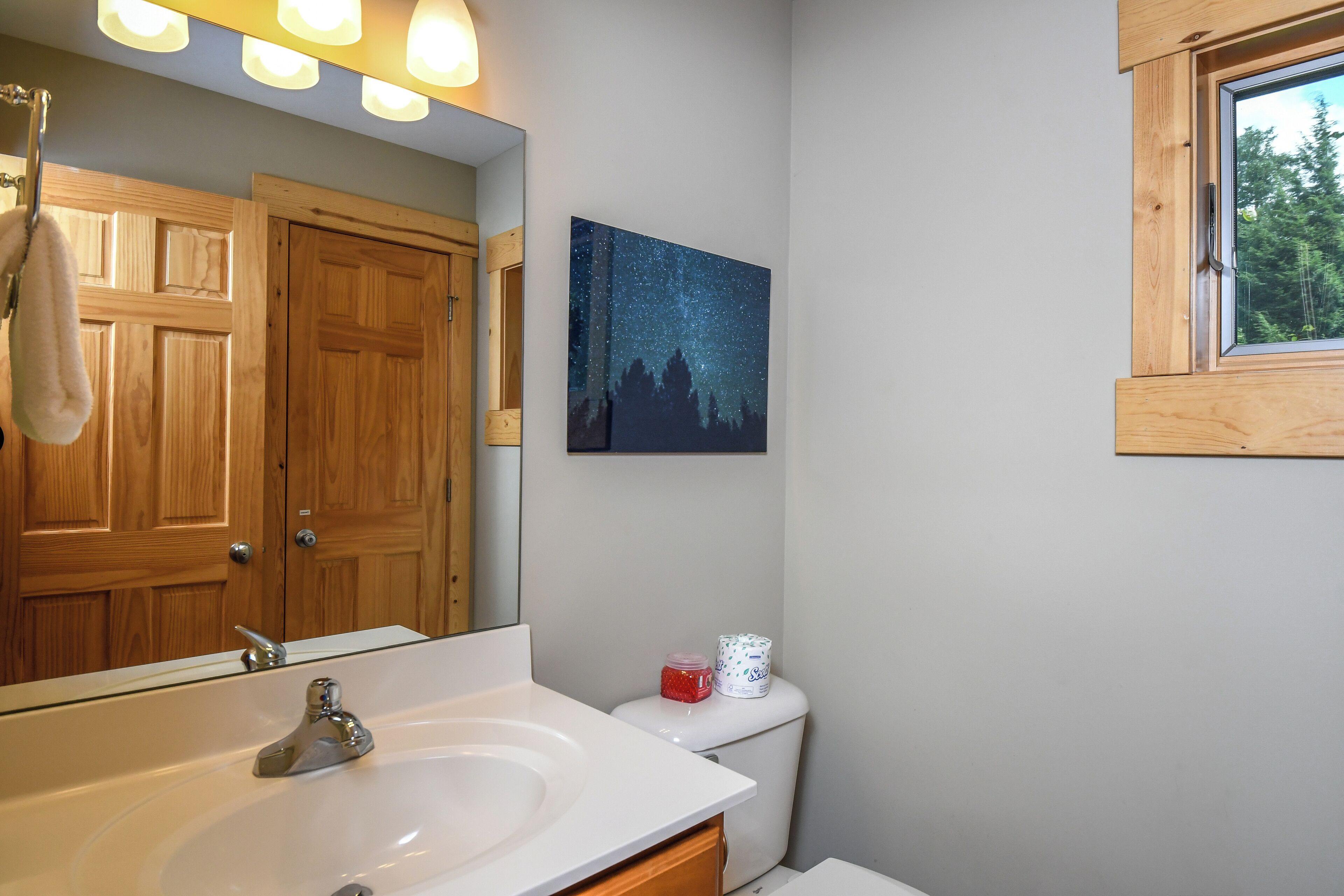 Un baño con puerta de madera, un lavamanos blanco, un inodoro y una pintura del cielo nocturno estrellado sobre el inodoro.