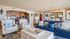 Ein geräumiges Wohnzimmer mit einem blauen Sofa, zwei Sesseln, einem Couchtisch und einer Küche im Hintergrund.
