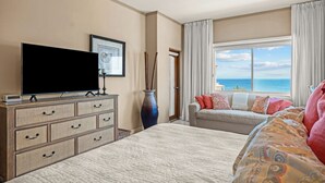 Ein Hotelzimmer mit Bett, Fernseher auf einer Kommode, Sofa mit Meerblick und einer Vase mit Zweigen.