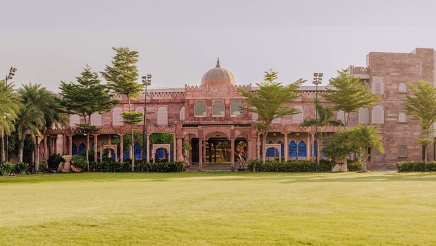 A large, historic building with a central dome and arched windows, surrounded by a well-maintained lawn and palm trees. - Amritam Palace (Jodhpur)