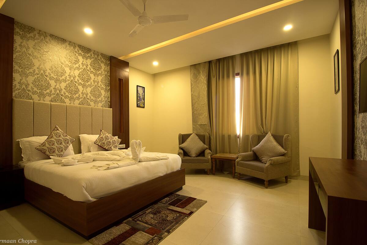 Quarto de hotel bem iluminado com uma cama grande, duas poltronas, uma mesinha e um ventilador de teto.