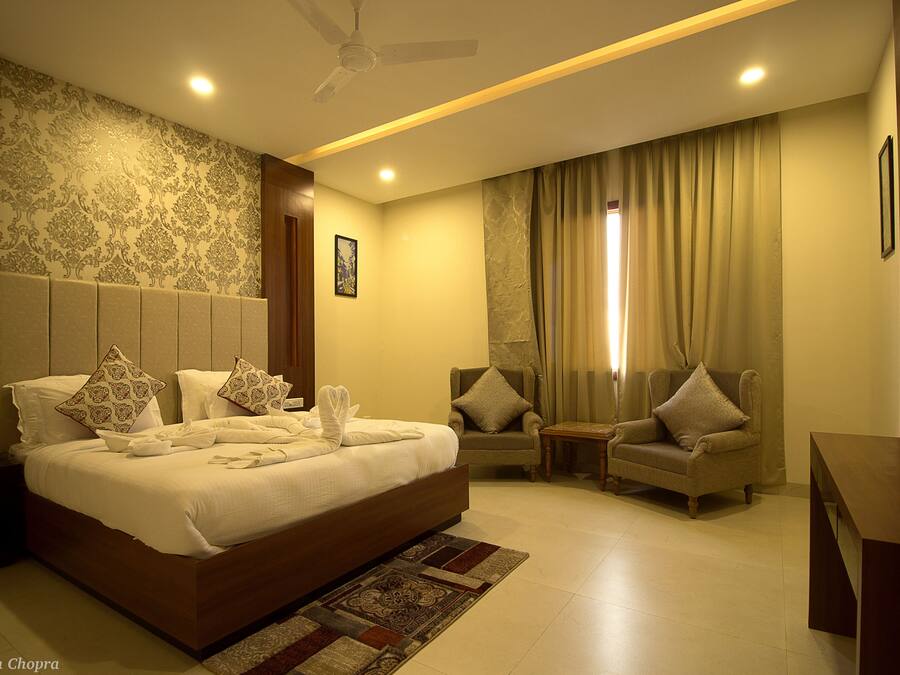 A well-lit hotel room with a large bed, two armchairs, a small table, and a ceiling fan.