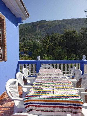 A balcony with a striped tablecloth, white chairs, and a view of green hills. - Gite Talassemtane, a cosy guesthouse in the middle of nature in Chefchaouen area (Chefchaouen)
