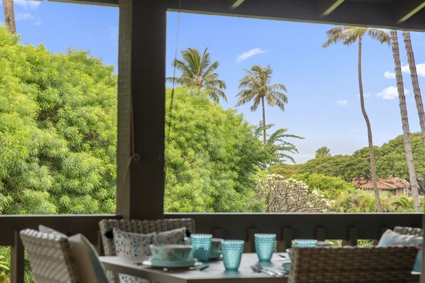 Un espace extérieur aménagé avec des meubles en osier et de la verrerie turquoise, offrant une vue sur un jardin luxuriant, avec des palmiers et un bâtiment situé au loin.