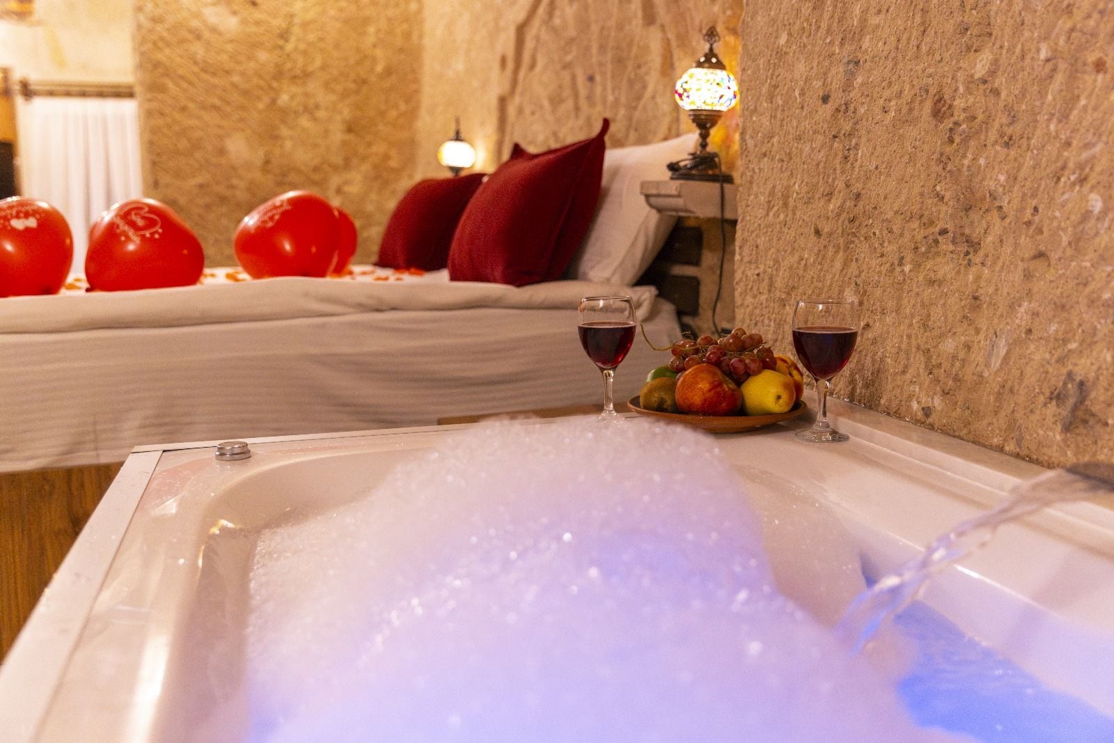 Una habitación de hotel con una bañera llena de burbujas, una cama con almohadas rojas y un frutero con uvas y manzanas.