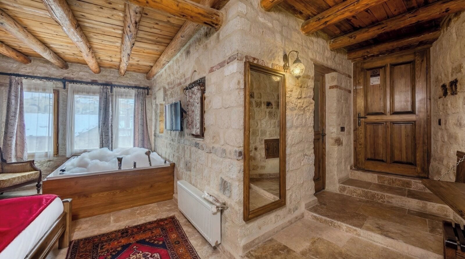A rustic room with a stone wall, wooden ceiling beams, a large mirror, a wooden door, and a bathtub with bubbles.