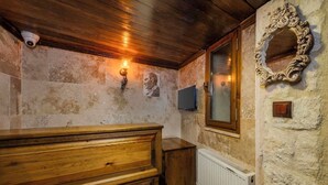 A rustic bathroom with a wooden bench, stone walls, a mirror, and a mounted television. - Zula Cave House (Ürgüp)