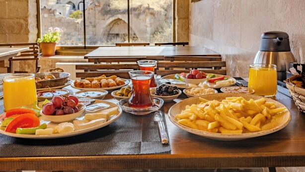 A breakfast setting with a variety of dishes, including fruits, pastries, and beverages, on a wooden table with a view of buildings through large windows. - Zula Cave House (Ürgüp)