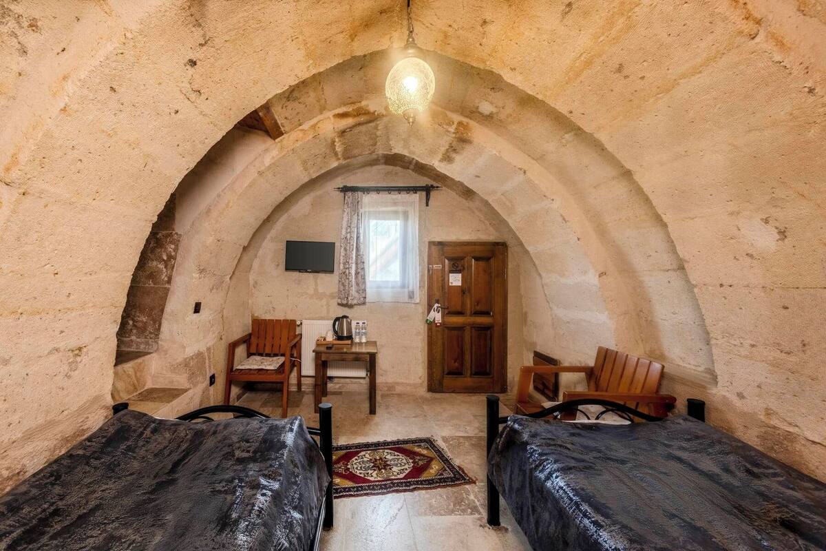 A cozy, rustic room with an arched ceiling, two wooden beds, a small table with chairs, a radiator, and a television on the wall.